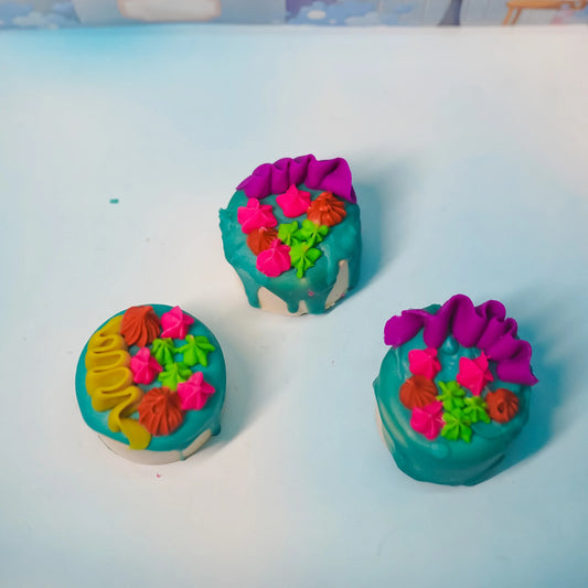 Three artisan bath bombs with teal icing and colorful floral decorations on a white surface