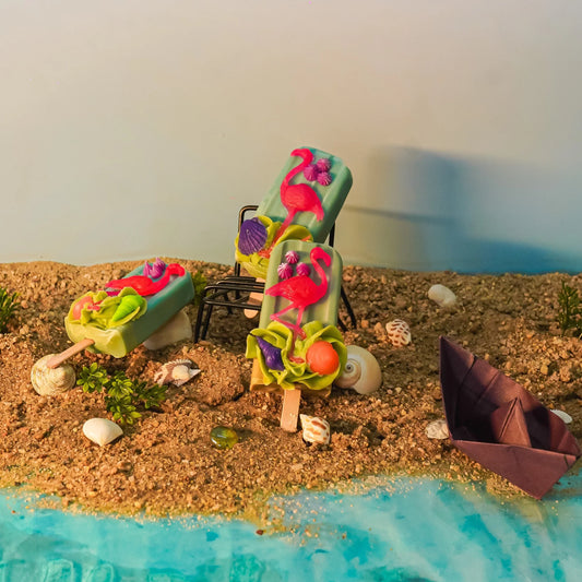 Artisan bath soaps shaped like popsicles with flamingo decor on beach sand, seashells, and paper boat