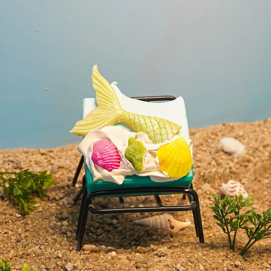Mermaid tail soap and colorful seashell soaps on a lounge chair in a sandy beach setting