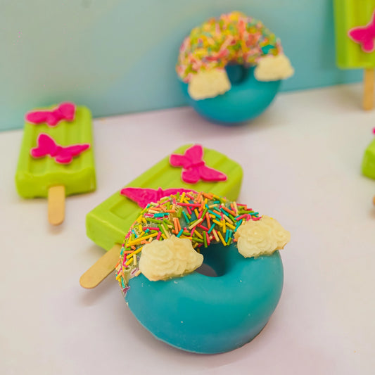 Colorful artisan bath products shaped like donuts and popsicles, decorated with sprinkles and butterfly designs.