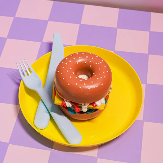 Whimsical donut-shaped burger bath bomb on yellow plate, pastel checkerboard background