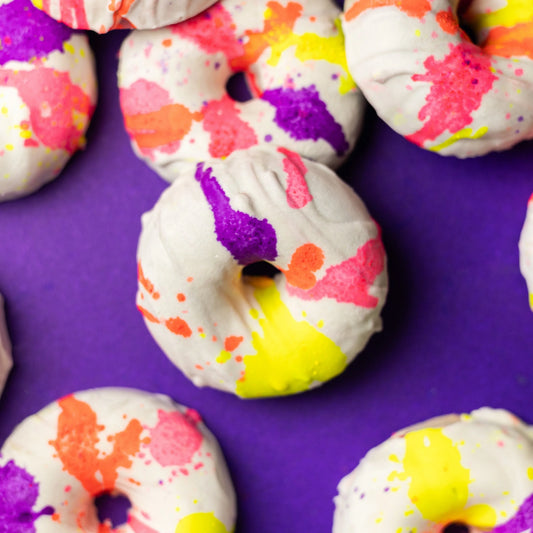 Handmade bath bombs shaped like donuts with colorful splatter designs on a purple background.