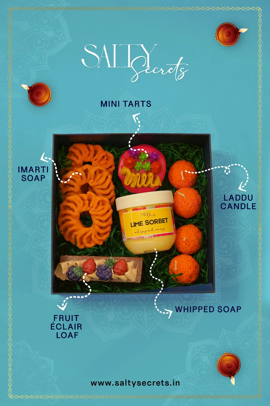 Diwali gift box with artisanal imarti soap, fruit éclair loaf, mini tarts, laddu candle, and whipped lime sorbet soap on a blue background.