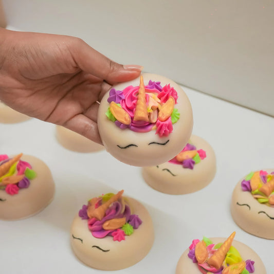 Hand holding round artisan soap with unicorn face design, colorful flowers, and a gold horn.