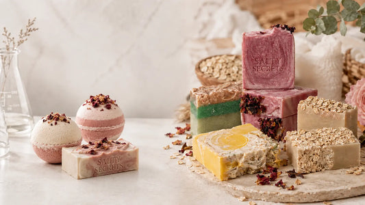 Realistic spa-style product photo of Salty Secrets handmade soaps and a bath bomb, featuring an engraved brown soap on a wooden tray, a green soap bar, and a white botanical bath bomb with soft flowers and cream fabric in the background.