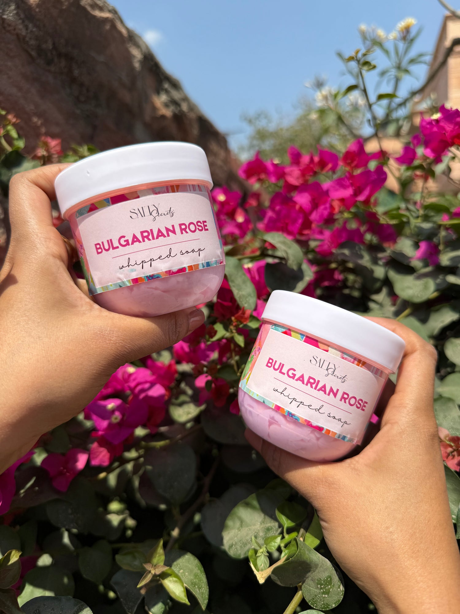 Two jars of Bulgarian Rose Whipped Soap being held in front of a vibrant floral background.